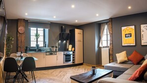 A modern living room with a sofa, a dining table with chairs, a kitchen area with appliances, and a large window.