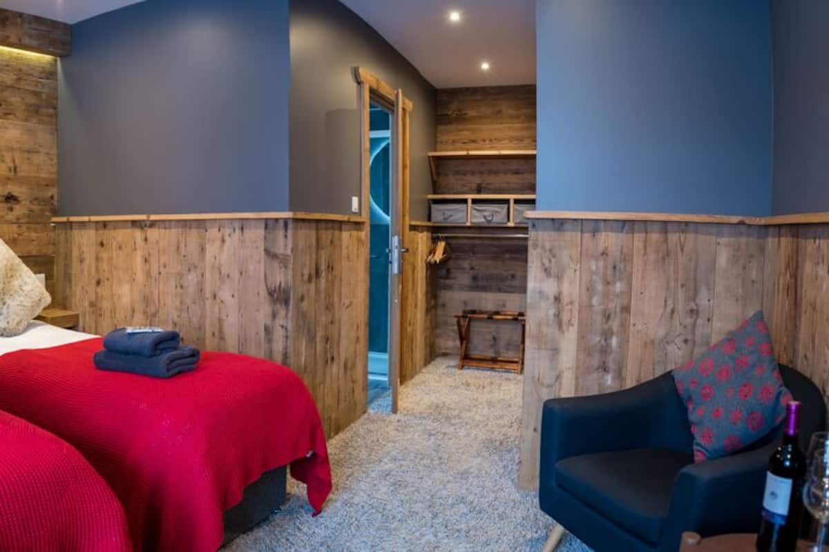 Habitación de hotel con dos camas, una pared de madera, un muro azul decorativo, una pequeña zona de estar y una escalera visible.