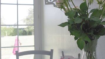 A table with a vase of pink roses, a white chair, and a window with a view of greenery.