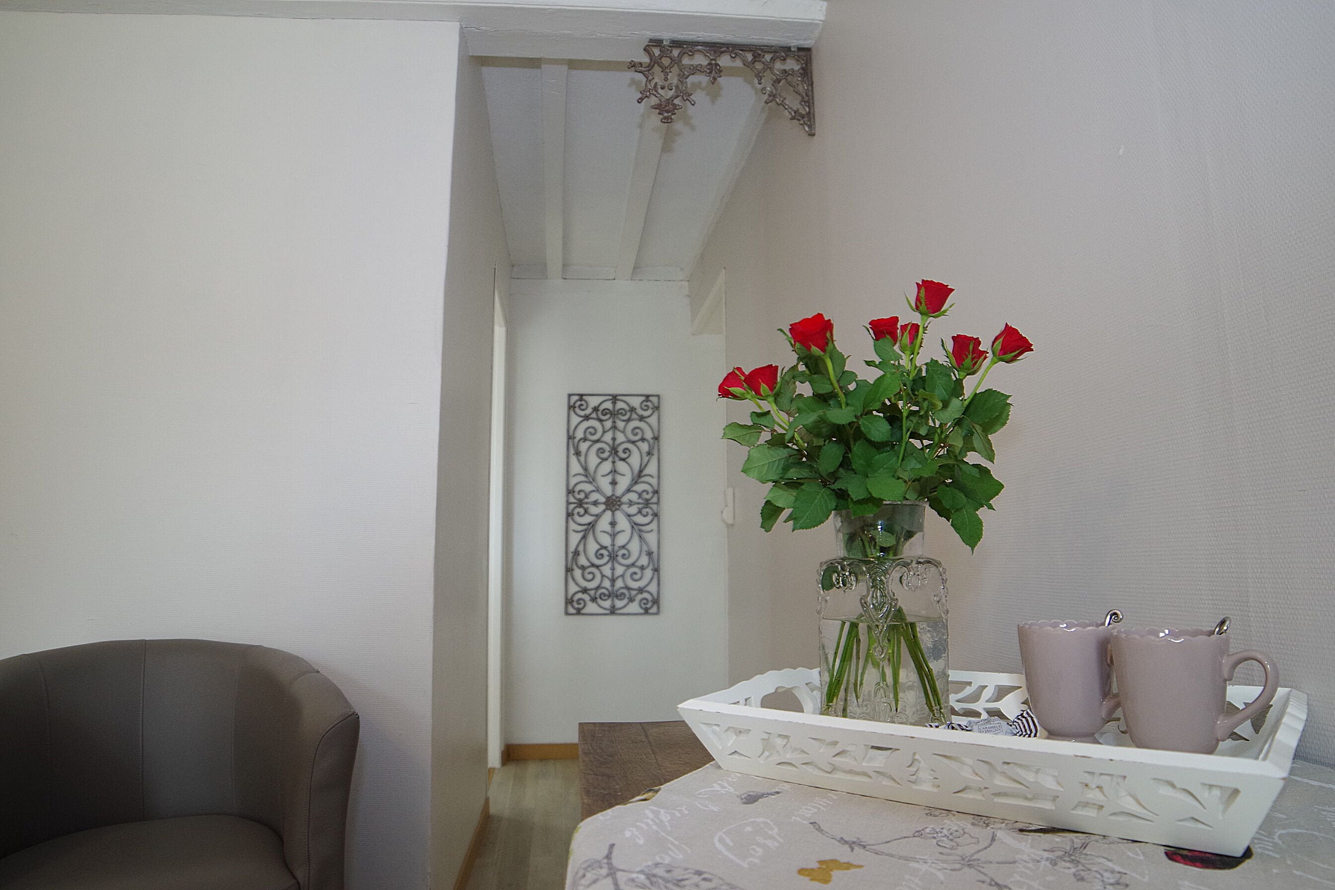 A room with a white wall, a decorative metal piece, a vase with red roses, and a tray with two mugs.