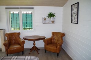 Une pièce comprenant deux fauteuils en cuir, une table ronde en bois, une fenêtre avec des barreaux en métal, une étagère fixée au mur ornée d’une plante, et une estampe botanique encadrée.