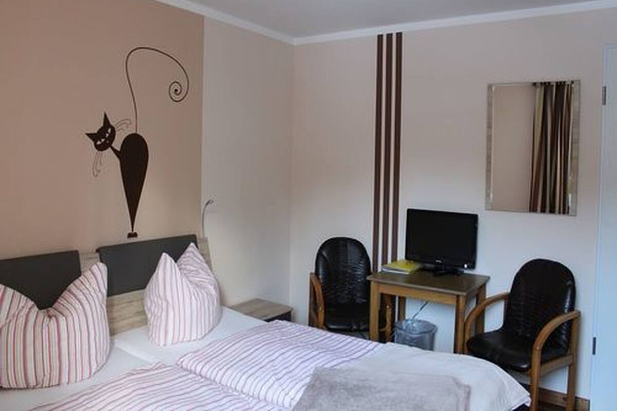 A bedroom with a bed, a desk with a computer, two chairs, and a wall-mounted TV.