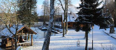 A rustic cabin with a snow-covered roof, surrounded by trees and a small dock.