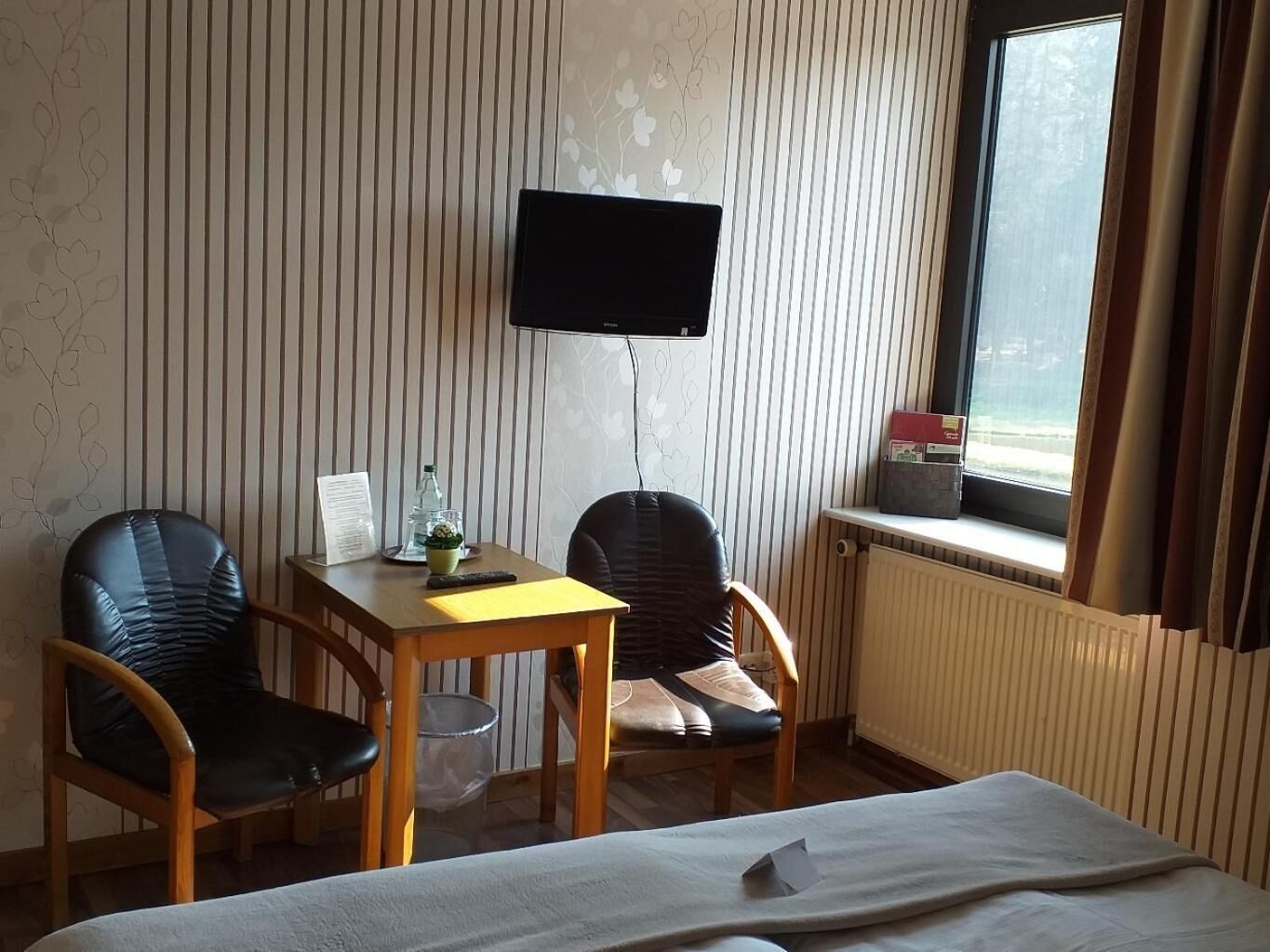 Habitación de hotel con cama, dos sillas, una mesita con una botella y un control remoto, un televisor montado en la pared y una ventana con cortinas.