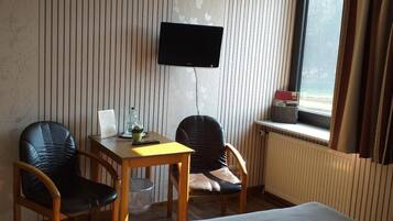 Habitación de hotel con cama, dos sillas, una mesita con una botella y un control remoto, un televisor montado en la pared y una ventana con cortinas.