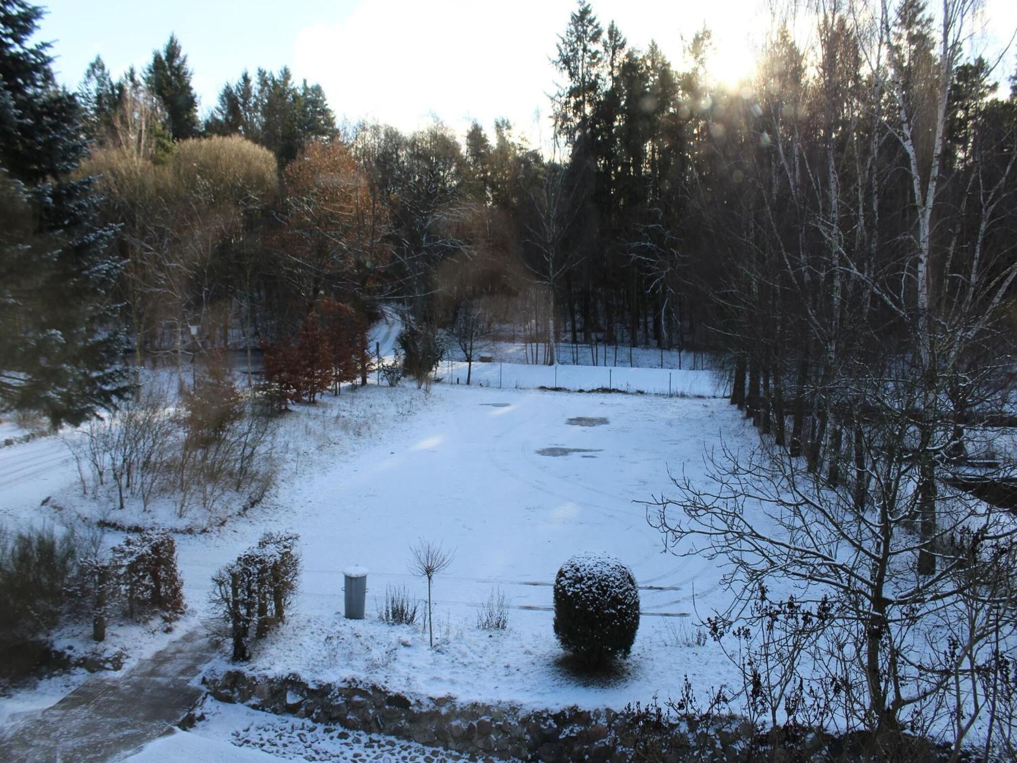 A snow-covered landscape with a stone wall, a small bush, and a pathway leading through a forest.