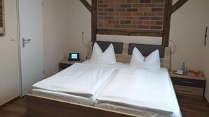 A double bed with white linens, a wooden headboard, a brick wall above, and a nightstand with a lamp and a small table. - Pension & Gasthaus Kattenstieg (Heiligengrabe)
