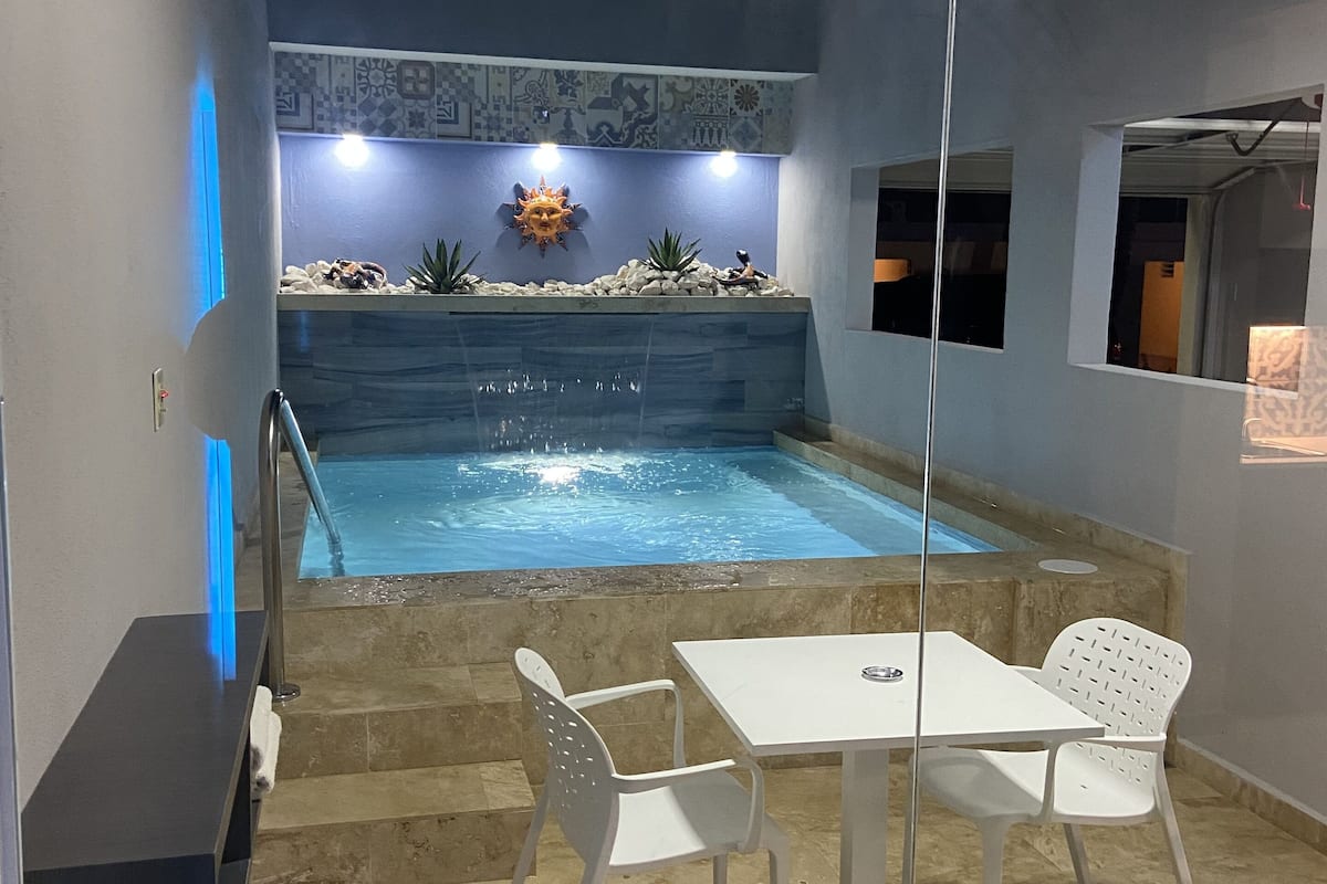 A small outdoor pool area with a white table and chairs, a decorative sun wall, and a tiled floor.