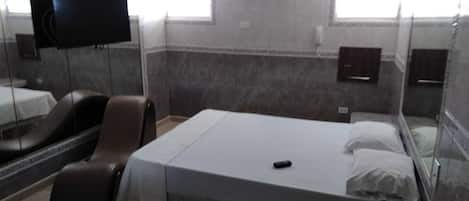 A hotel room with a bed, a TV, a chair, and a small bathroom.