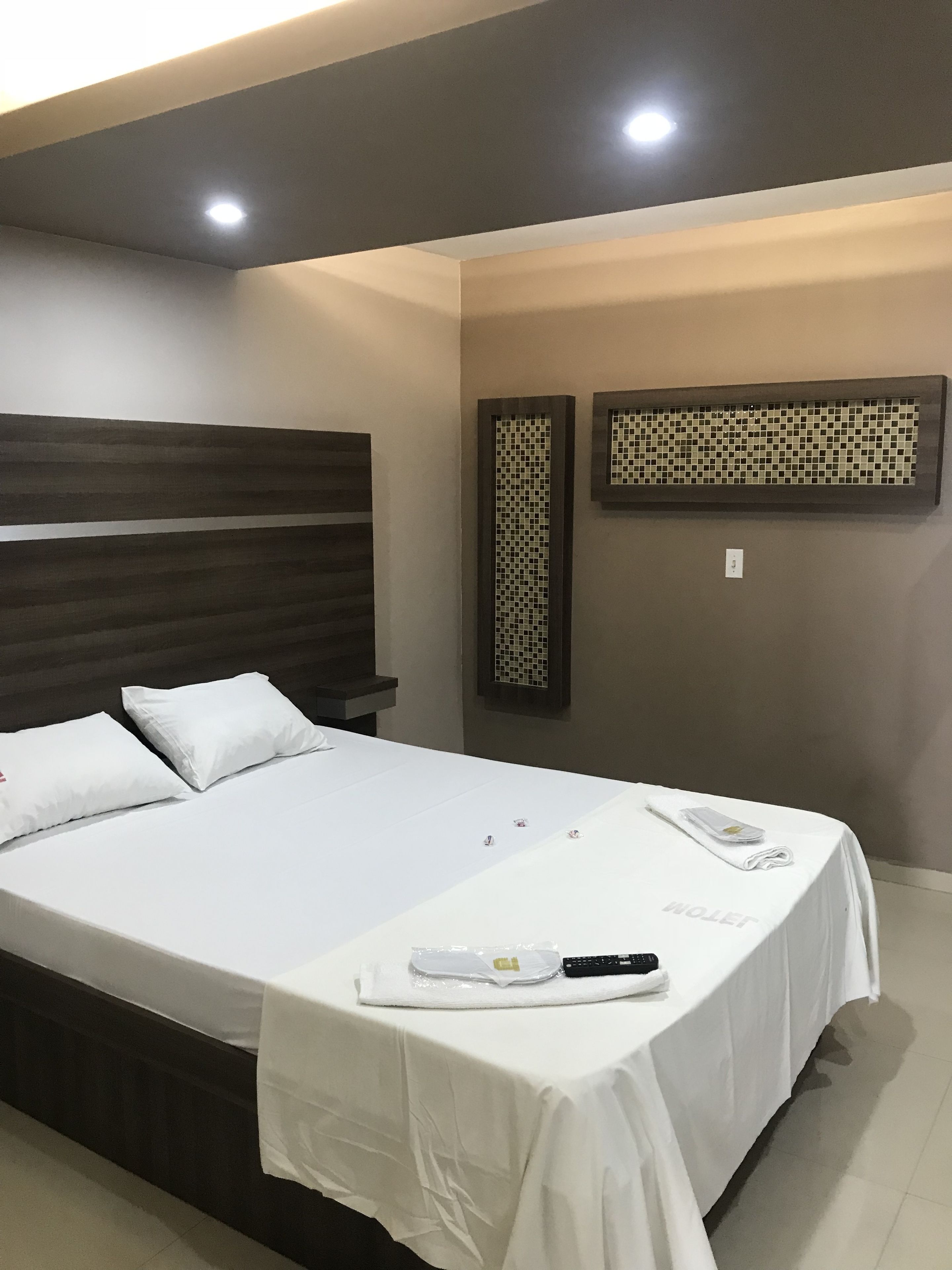 A hotel room with a large bed, a headboard, a wall-mounted air conditioning unit, and a small table with a remote control and a phone.