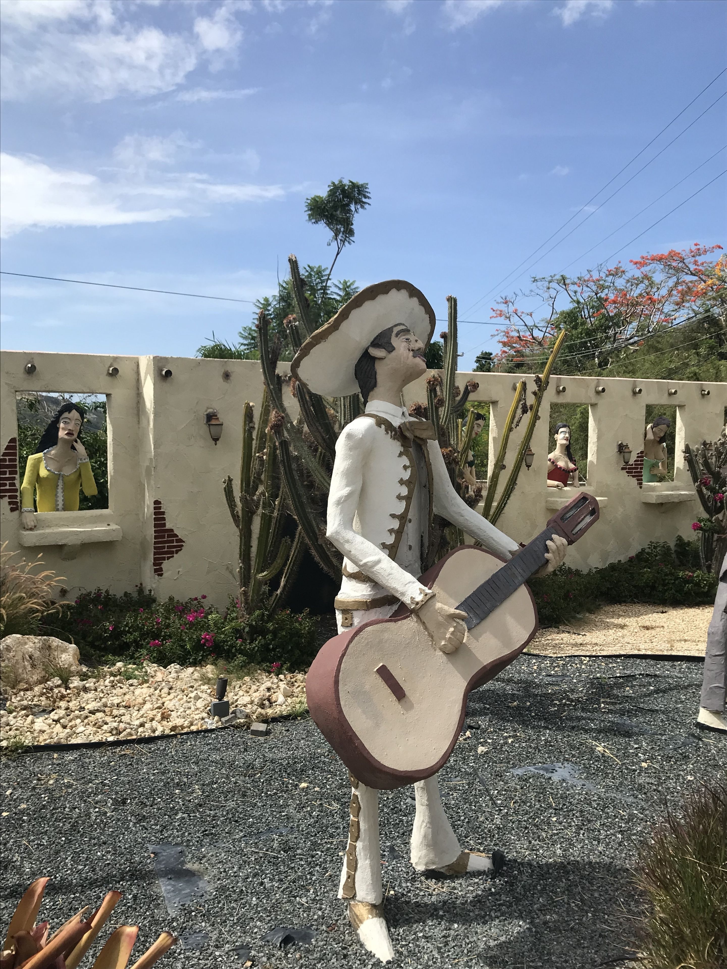 A statue of a man in a white outfit playing a guitar, set against a wall with cacti and a clear sky.