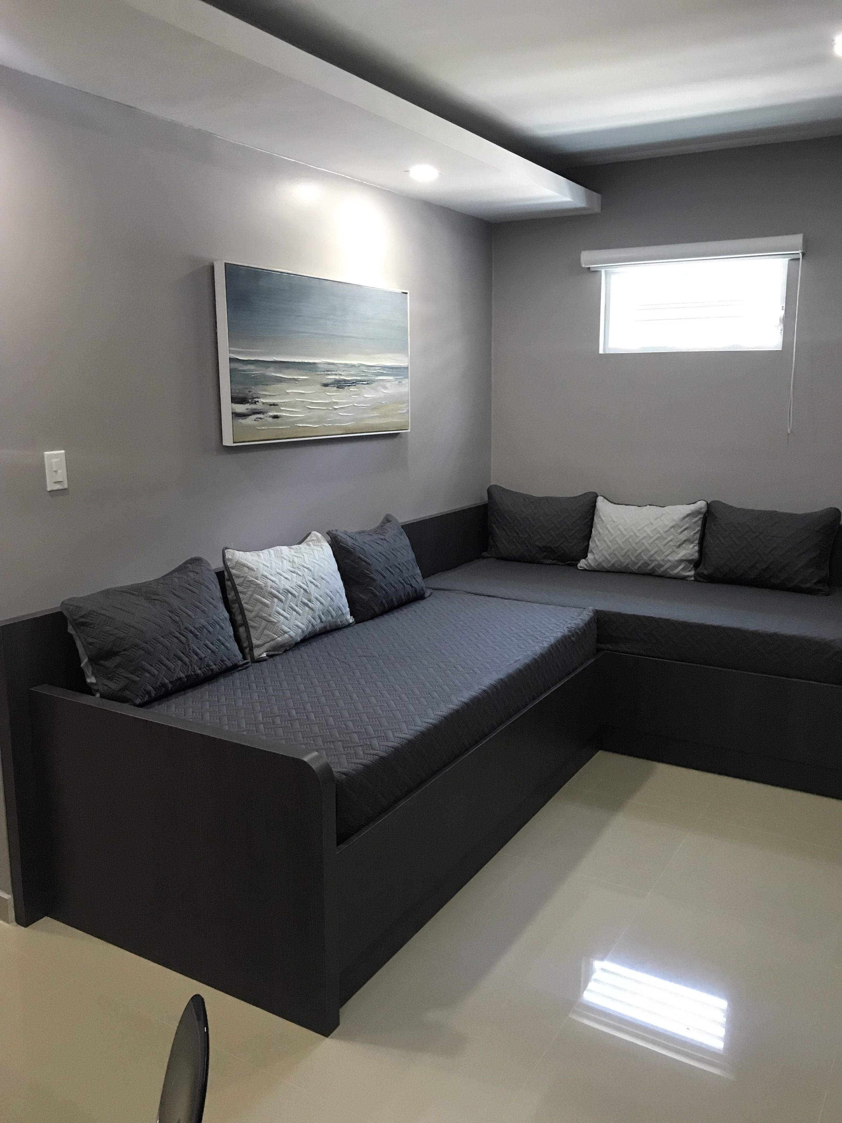 Un sofá seccional moderno en forma de L con tela gris acolchada y múltiples cojines, en una habitación contemporánea con un cuadro grande de una escena de playa, una ventana con persianas y una luz empotrada en el techo.