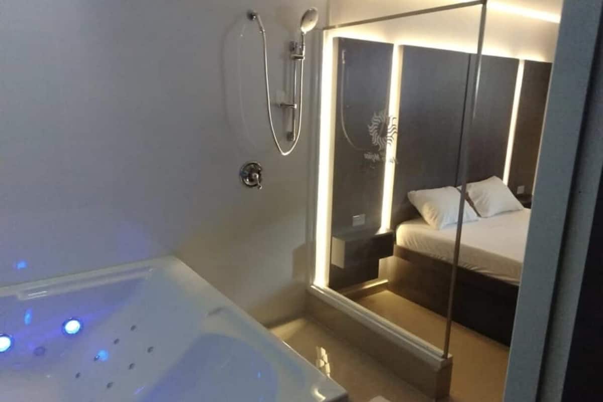 A modern bathroom with a large bathtub, a shower, and a mirror reflecting a bed with white bedding.