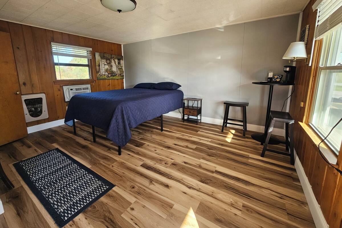 A bedroom with a bed, a small table, a lamp, a heater, and a window.