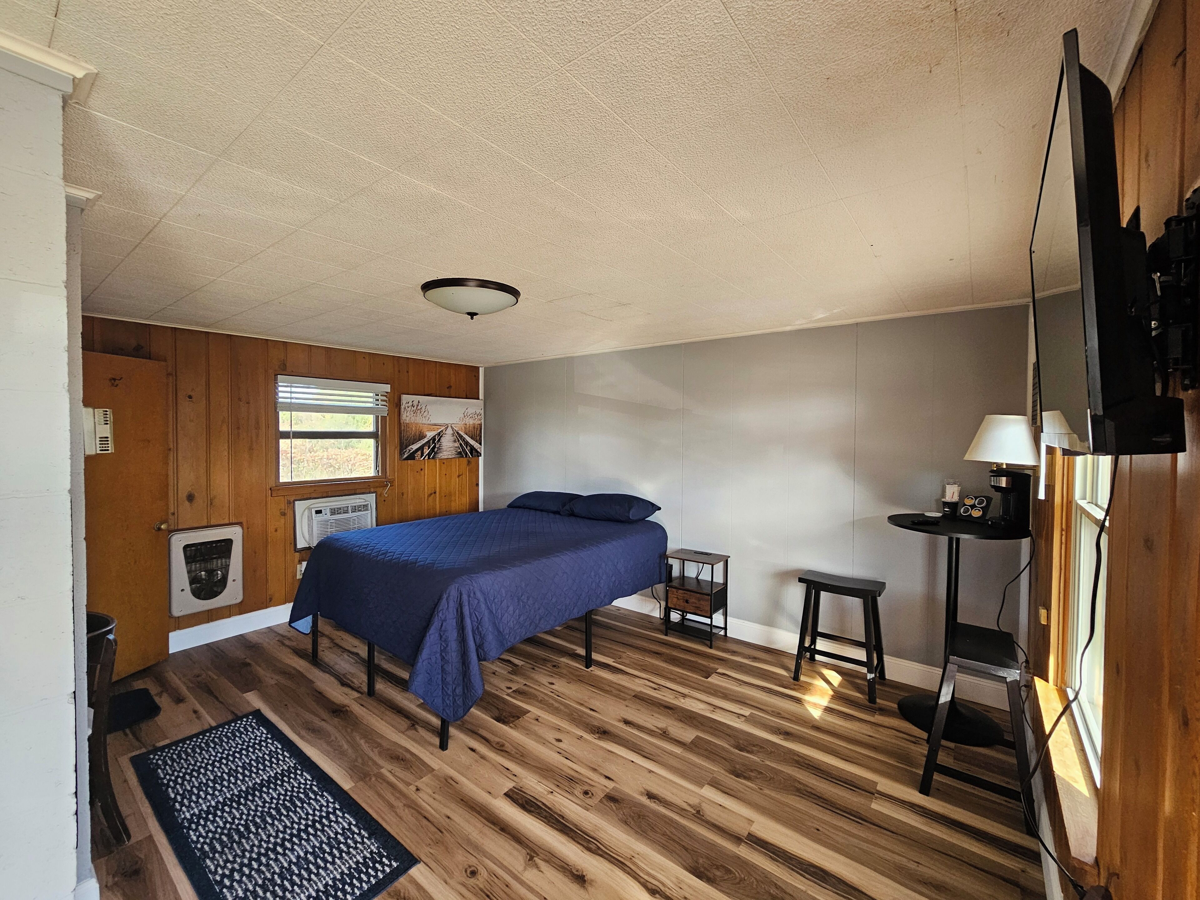 Une chambre avec un plancher en bois, un lit recouvert d’une courtepointe bleue, une petite table de chevet avec une lampe et un téléviseur fixé au mur.