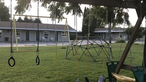 A playground with swings, a climbing structure, and a building in the background. - The Last Resort (Hart)