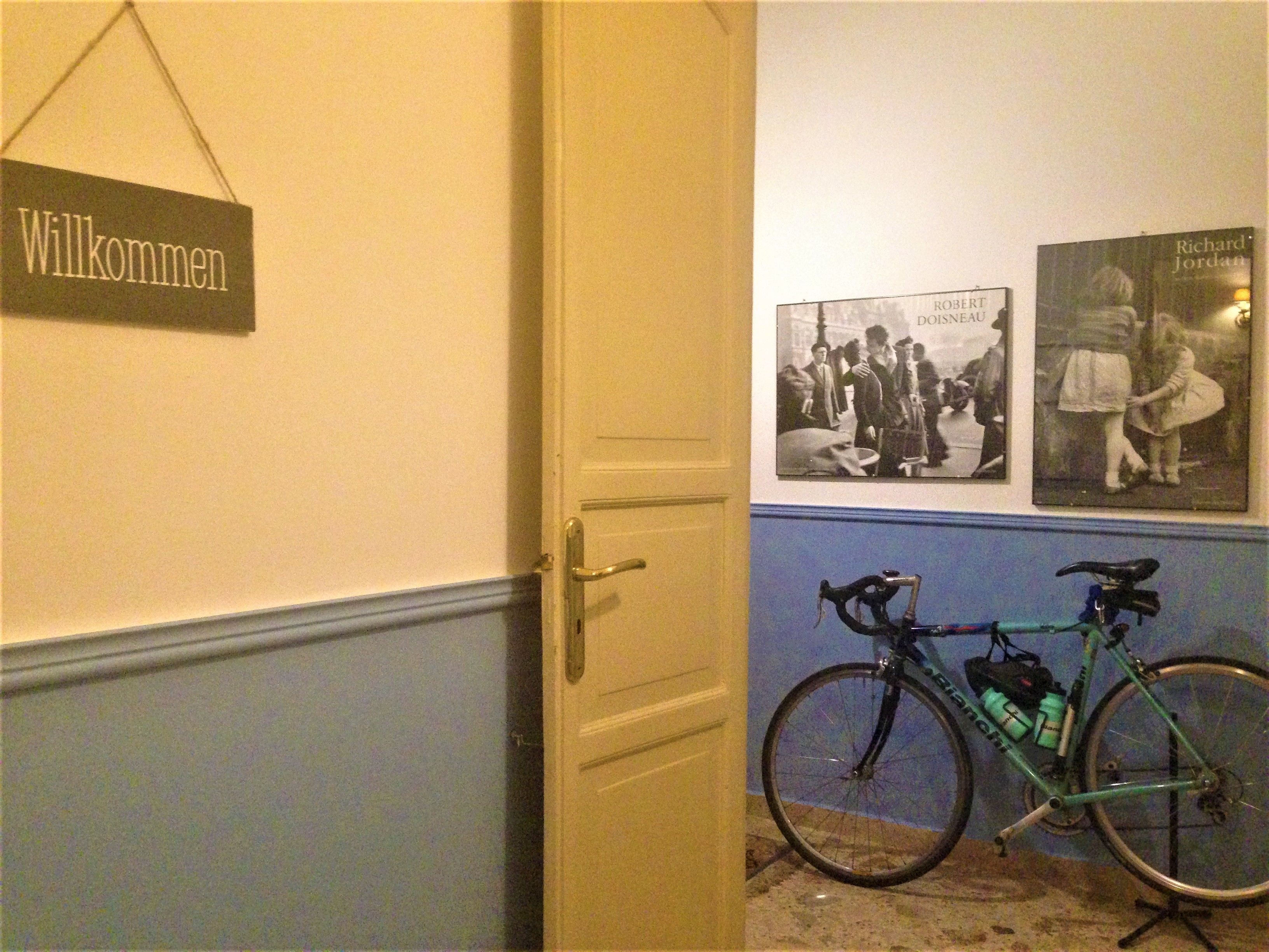 Een gang met een 'Willkommen' bord, een fiets en twee ingelijste foto's aan de muur.