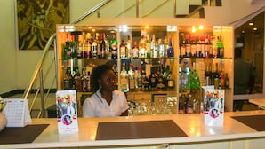 A bar area with a woman behind the counter, shelves stocked with various bottles, and a staircase in the background. - New Rivoli Hotel Benin (Cotonou)