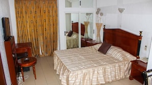A bedroom with a bed, a desk, a chair, a mirror, and a window with curtains. - New Rivoli Hotel Benin (Cotonou)