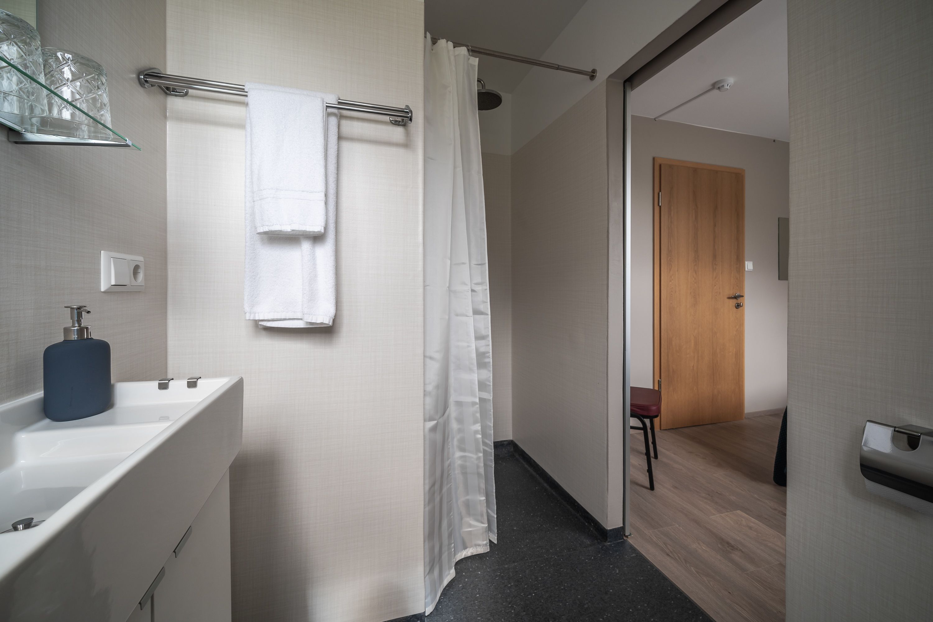 Un baño moderno con un lavamanos blanco, cabina de ducha con mampara de vidrio y una puerta de madera que da a otra habitación.