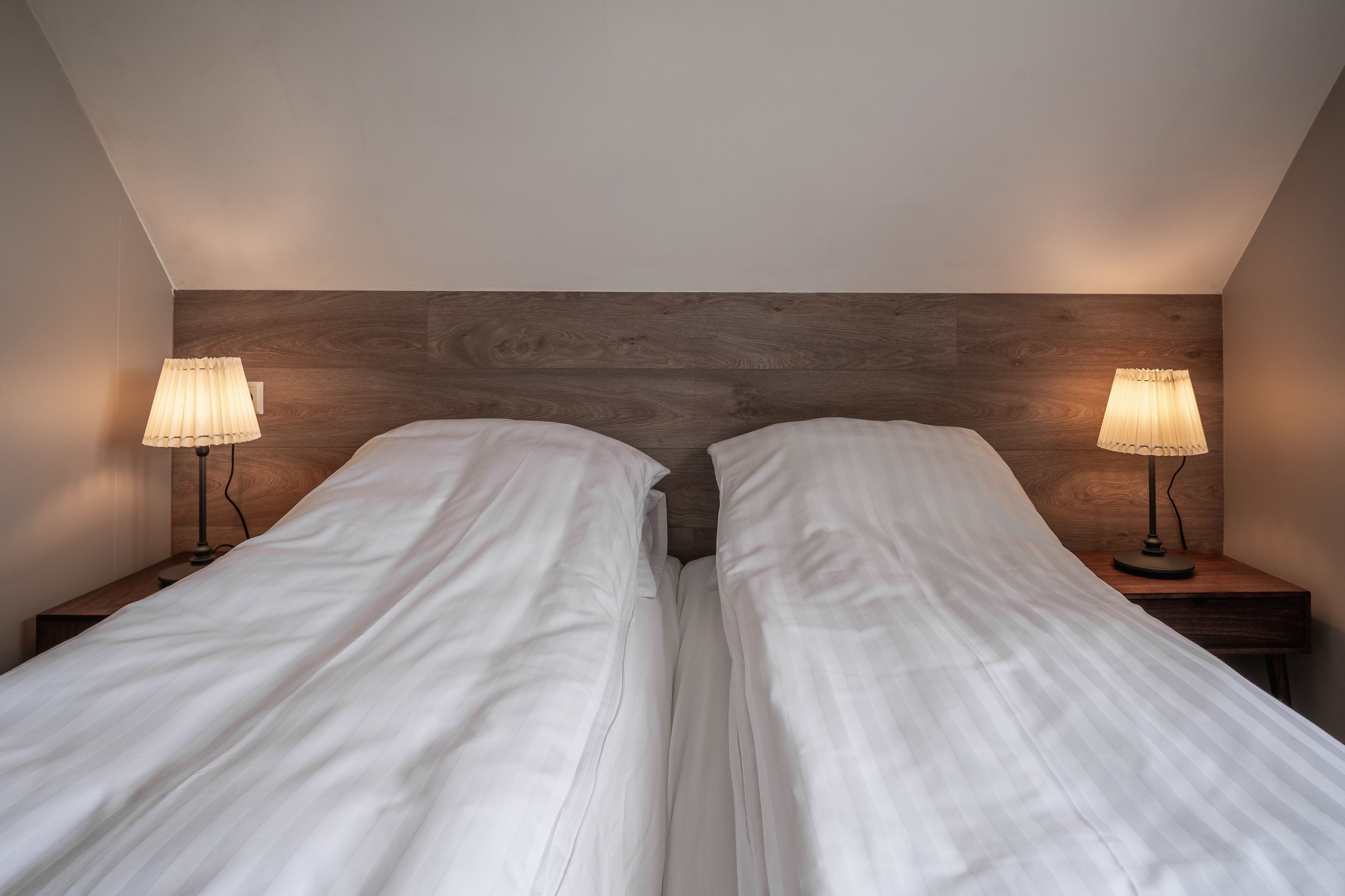 Uma cama de casal com roupa de cama branca em quarto com cabeceira de madeira e duas luminárias de cabeceira.