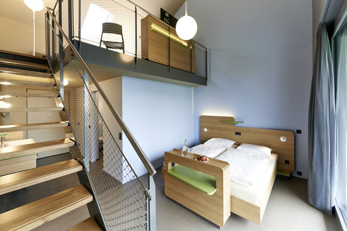Una habitación de hotel moderna con una escalera de madera, una cama con mesita de noche verde y vistas a la ciudad.
