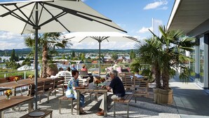 An outdoor seating area with tables and umbrellas, surrounded by palm trees and a view of a town.