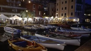A marina with several boats docked, a crowd of people, and buildings in the background.