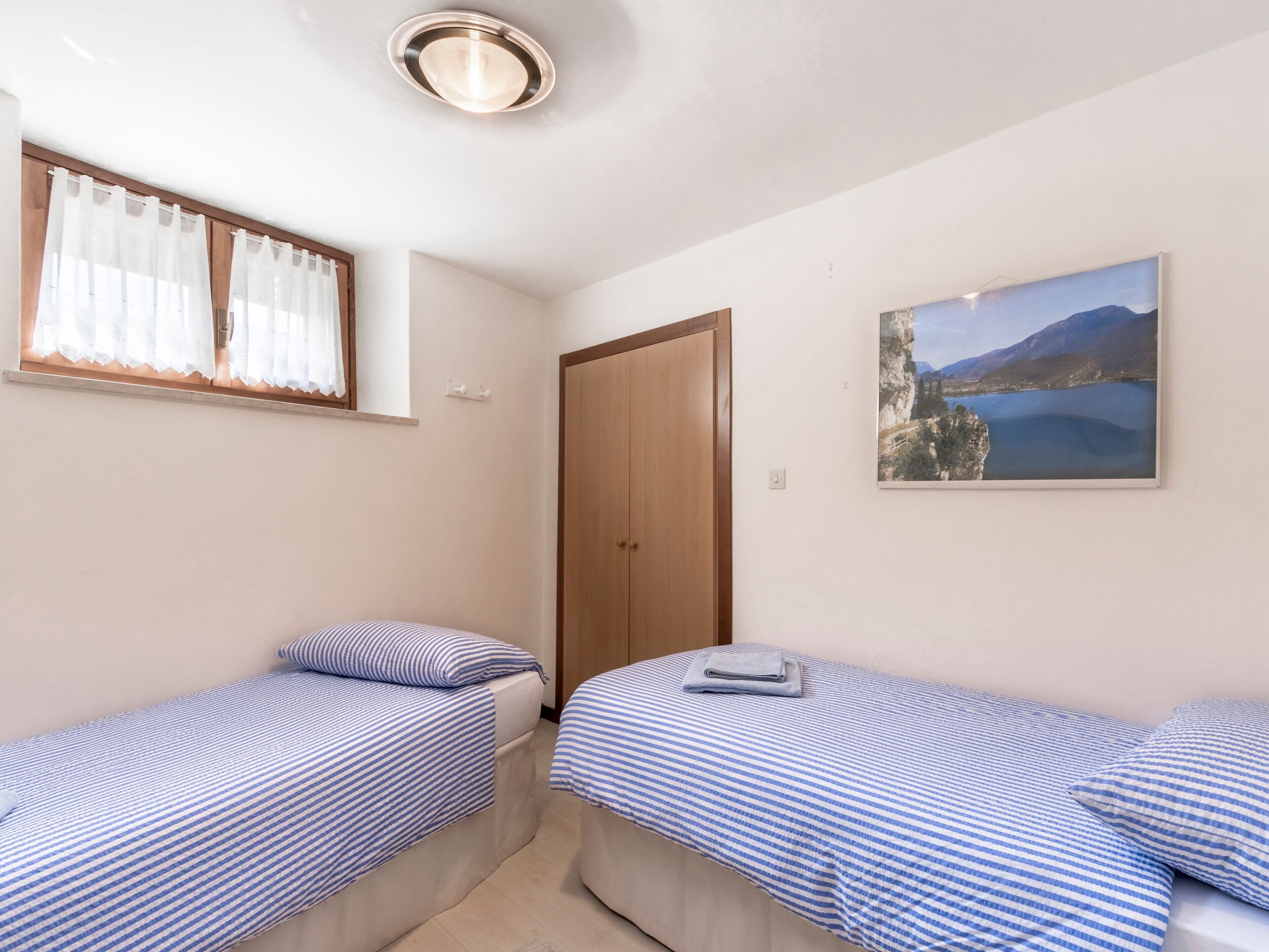 Un dormitorio con dos camas individuales, un armario de madera, una ventana con cortinas traslúcidas y un cuadro de paisaje en la pared.