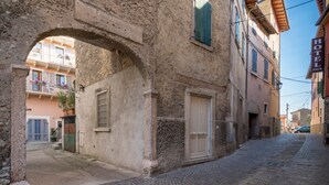 Une ruelle étroite pavée, surmontée d’une arche en pierre, un bâtiment portant une enseigne indiquant « HÔTEL PACE », et un ciel d’un bleu limpide.