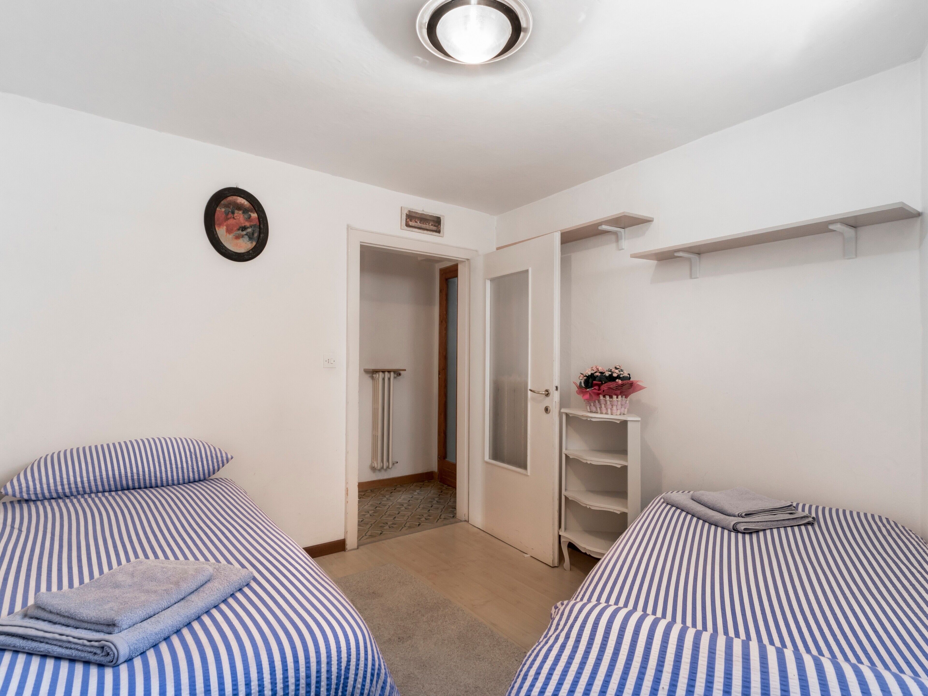 Habitació amb dos llits, un radiador, una porta que dona al bany i una estant amb un ram de flors.