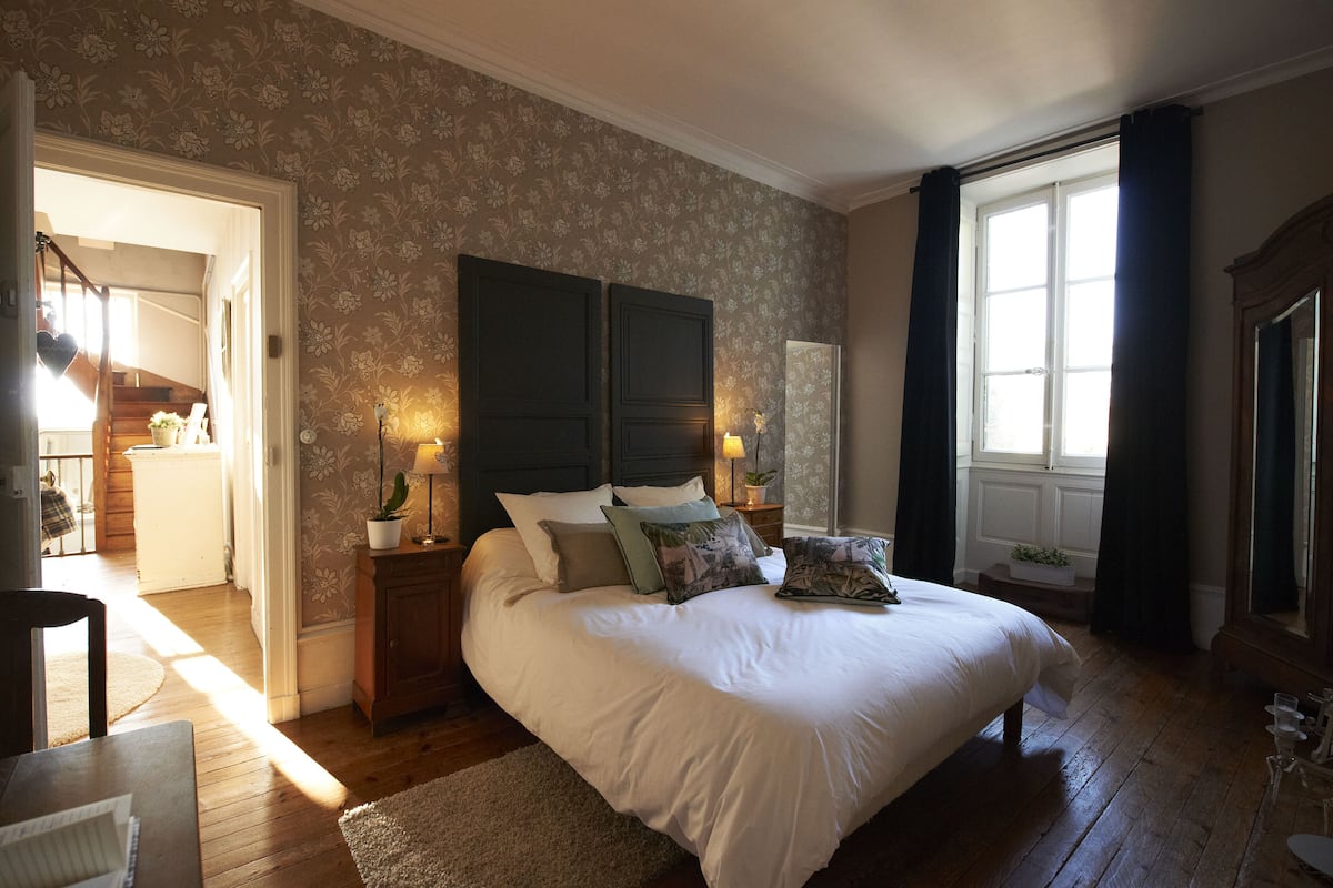 A bedroom with a large bed, wooden floors, a floral wallpaper, and a view of the outside through a window.