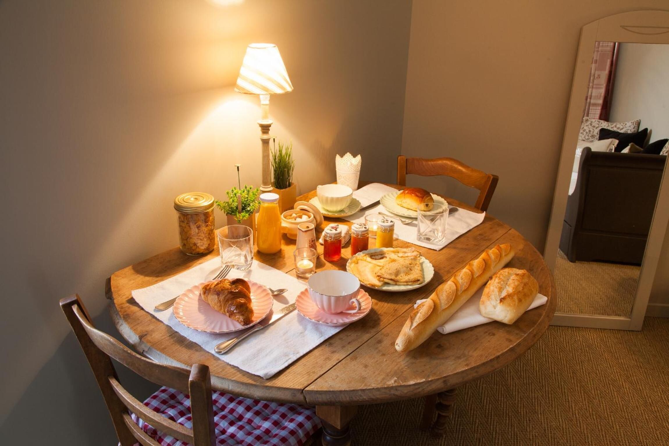 A wooden dining table with a lamp, various food items, and a chair with a checkered cushion.
