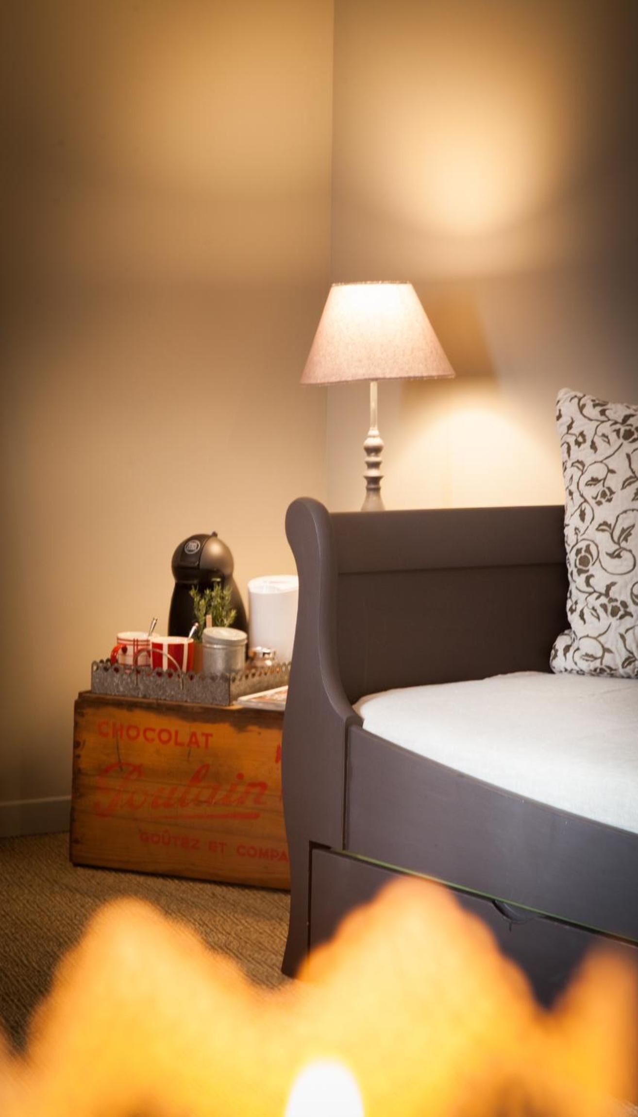 A bedroom with a bed, a lamp, a nightstand with a decorative item, and a box labeled 'CHOCOLAT'.