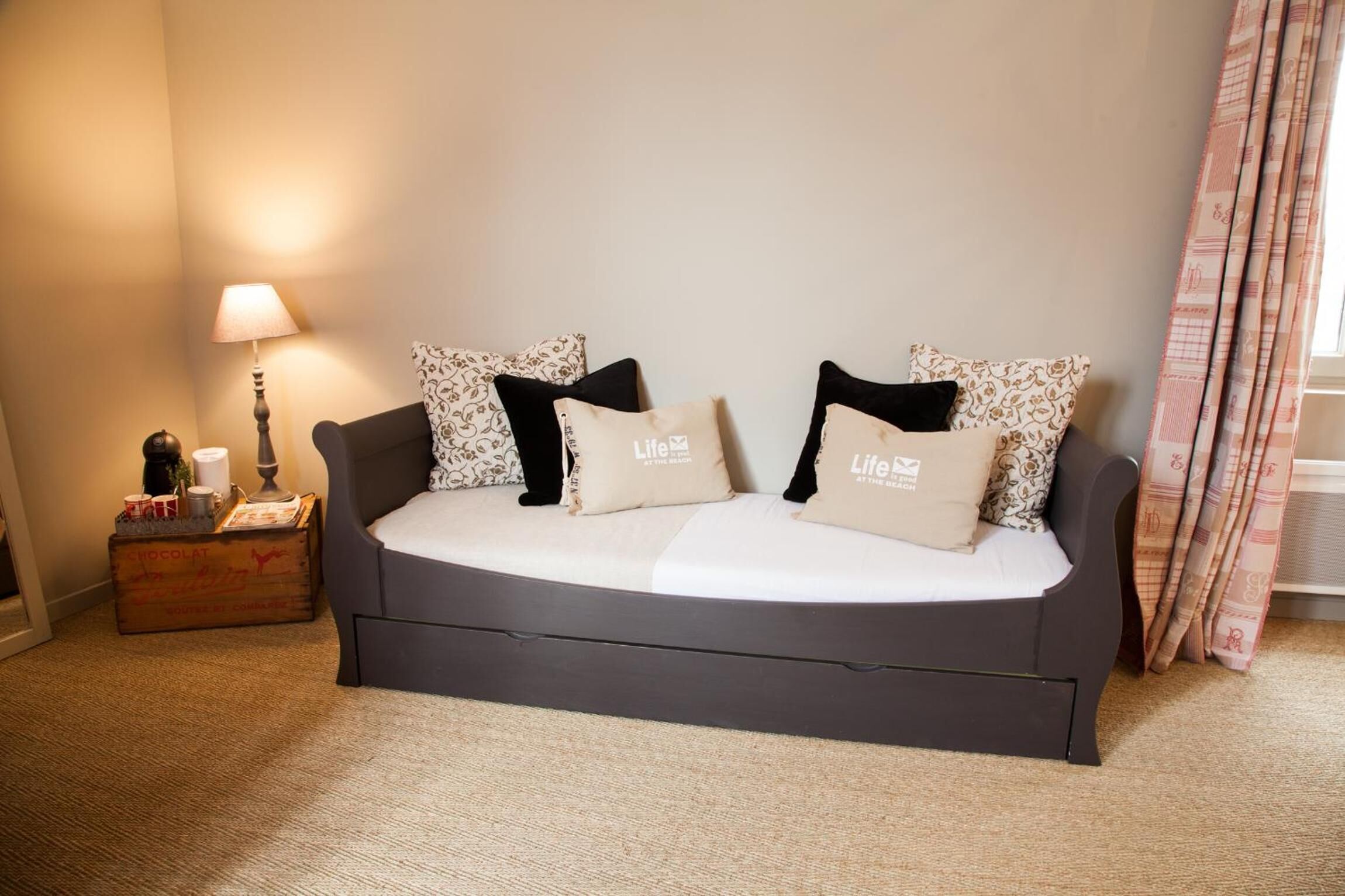 Un sofá de color oscuro con cojines estampados y dos almohadas de la marca 'Life', una lámpara y un pequeño cofre de madera con objetos encima.