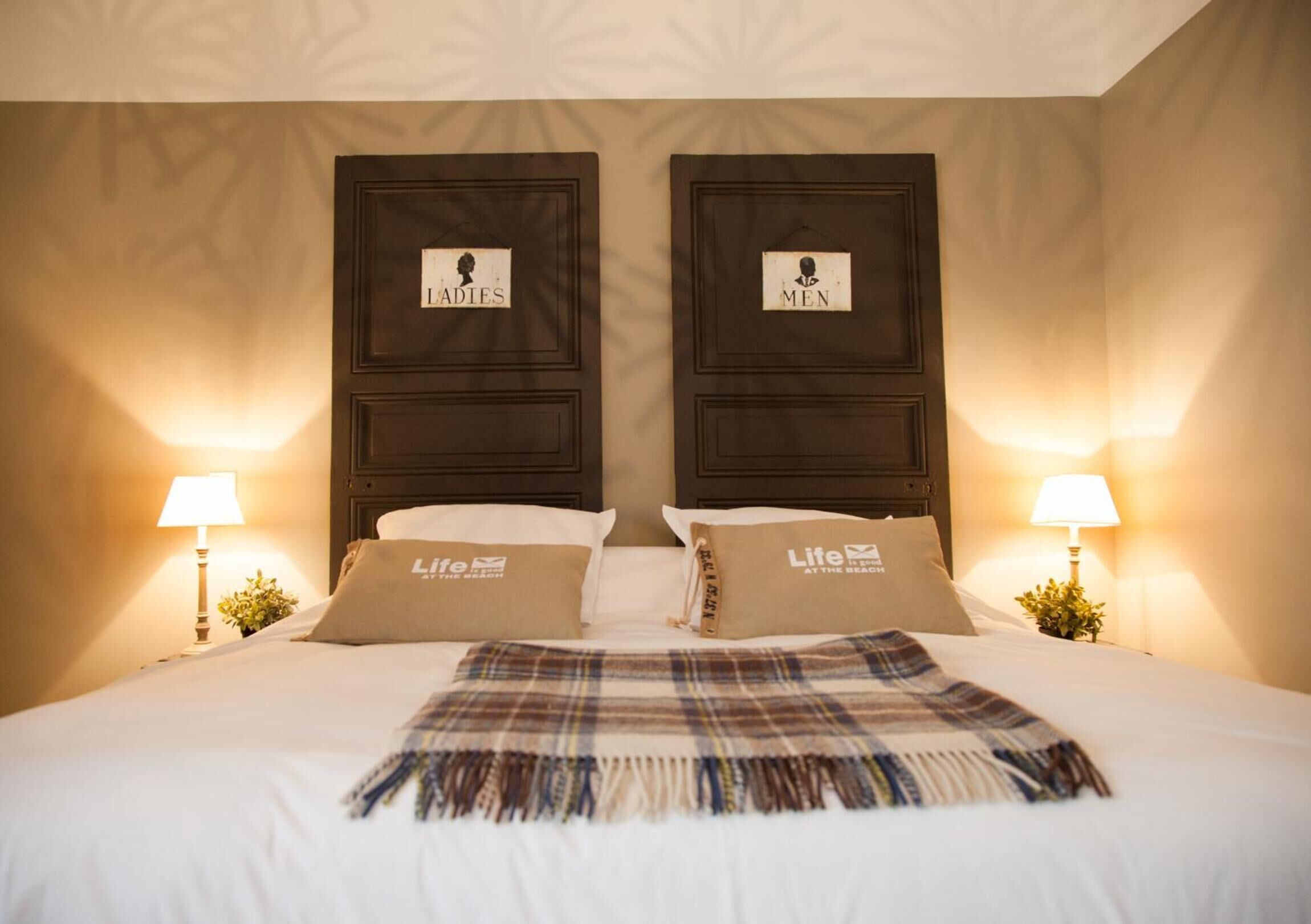 Une chambre d’hôtel avec un grand lit, deux tables de chevet avec des lampes et deux têtes de lit portant les inscriptions « Dames » et « Messieurs ».