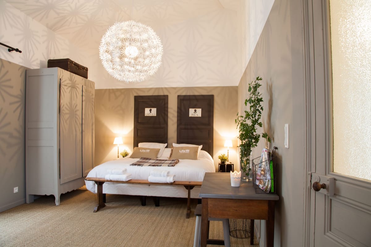 A bedroom with a bed, bedside tables, a desk, a hanging light fixture, and a wardrobe.