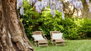 Dos sillones de madera con cojines estampados en un jardín con césped, un tronco de árbol grande y glicinias moradas colgando arriba.