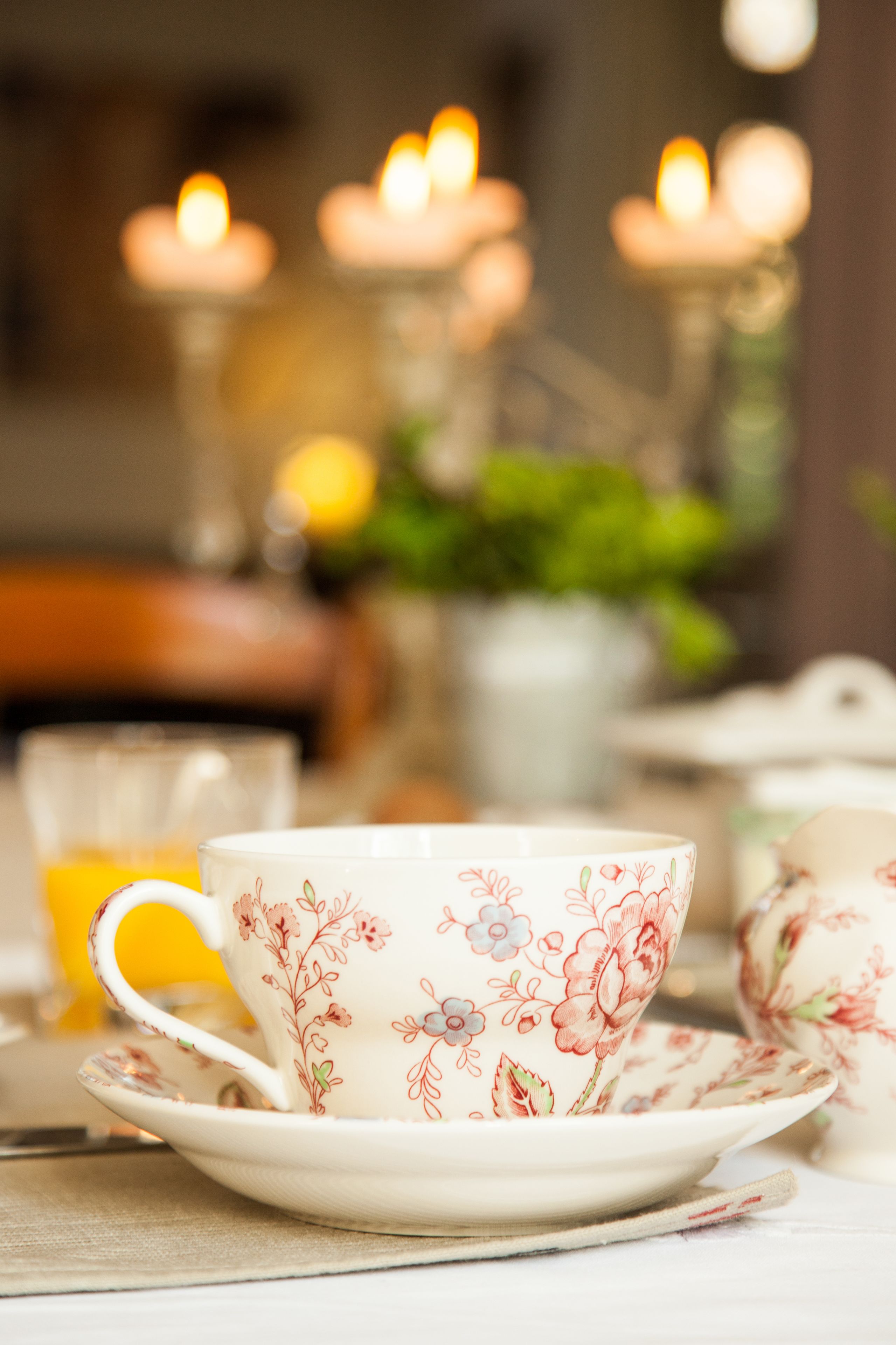 A floral-patterned tea set with a cup and saucer, a glass of orange juice, and a candle in the background.