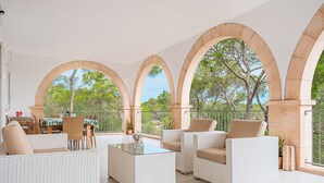 A spacious patio with white wicker furniture, a glass coffee table, and a view of trees through arched openings. - Villa Rosa – Cala Mondragó (Santanyí)