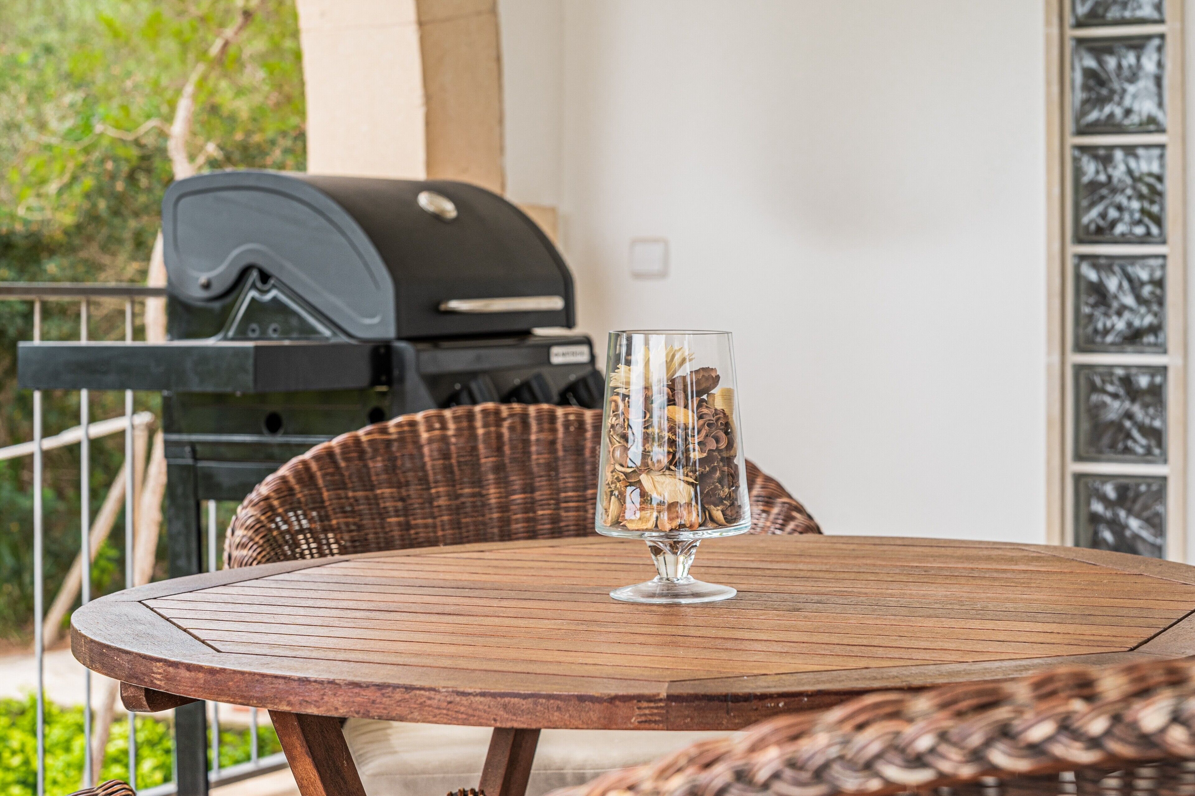 A round wooden table with a clear glass vase, a wicker chair, and a barbecue grill in the background.