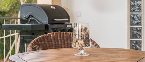 A round wooden table with a clear glass vase, a wicker chair, and a barbecue grill in the background.