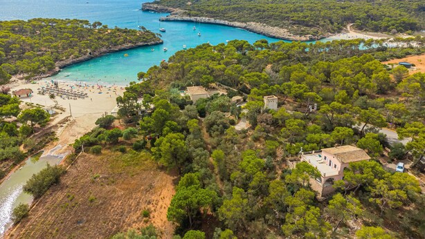 An aerial view of a coastal area with a beach, boats, and a small building surrounded by trees. - Villa Rosa – Cala Mondragó (Santanyí)