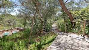 A stone pathway leading through a wooded area with trees and a stone bench area. - Villa Rosa – Cala Mondragó (Santanyí)