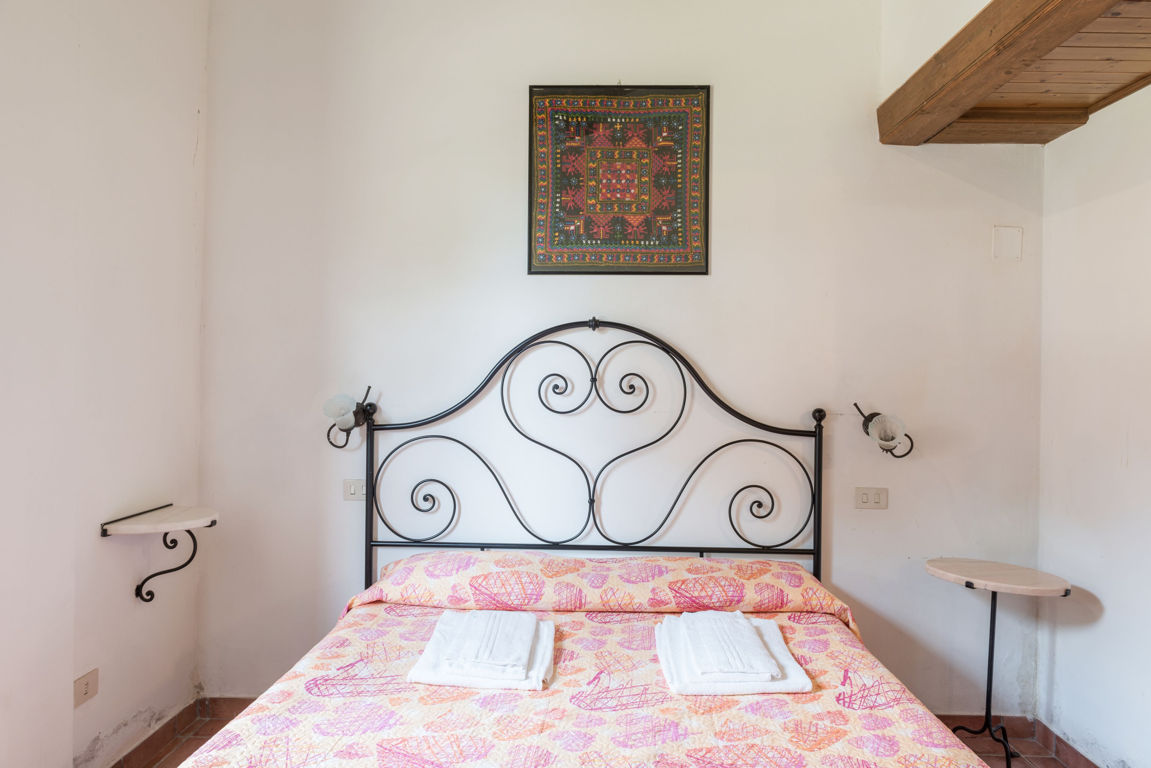 Una habitación con estructura de cama metálica, colcha estampada, una mesita pequeña y un adorno colgante en la pared.