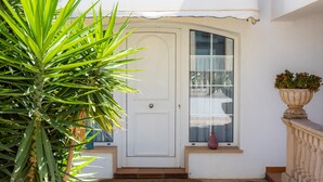 A white door with a glass window, a potted plant to the left, and a small window with a curtain to the right. - Central Holiday Home Near the Beach – Villa Amoros (Cala Ratjada)