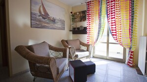 Una stanza con due poltrone in vimini, un poggiapiedi e un quadro raffigurante una barca a vela, con tende dai colori vivaci.