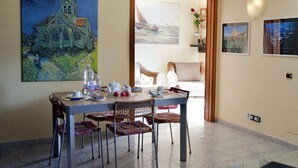 Un'area pranzo con un tavolo apparecchiato per due, sedie, un quadro raffigurante una chiesa e vista mare attraverso una porta.