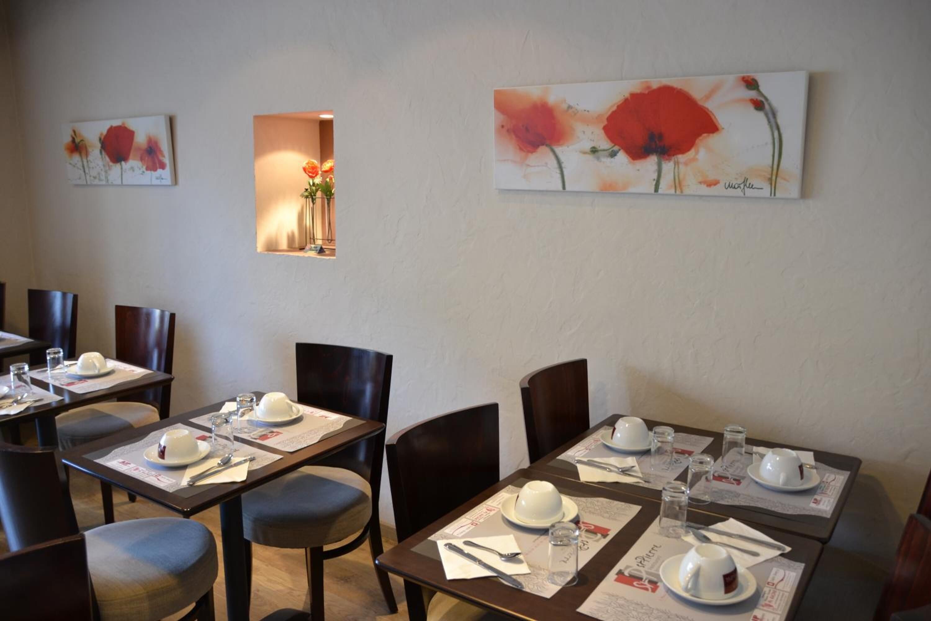 Un comedor con mesas dispuestas para dos comensales, cada una con un plato blanco, una taza blanca, cubiertos y una copa. En las paredes hay pinturas florales y una pequeña ventana adornada con flores.