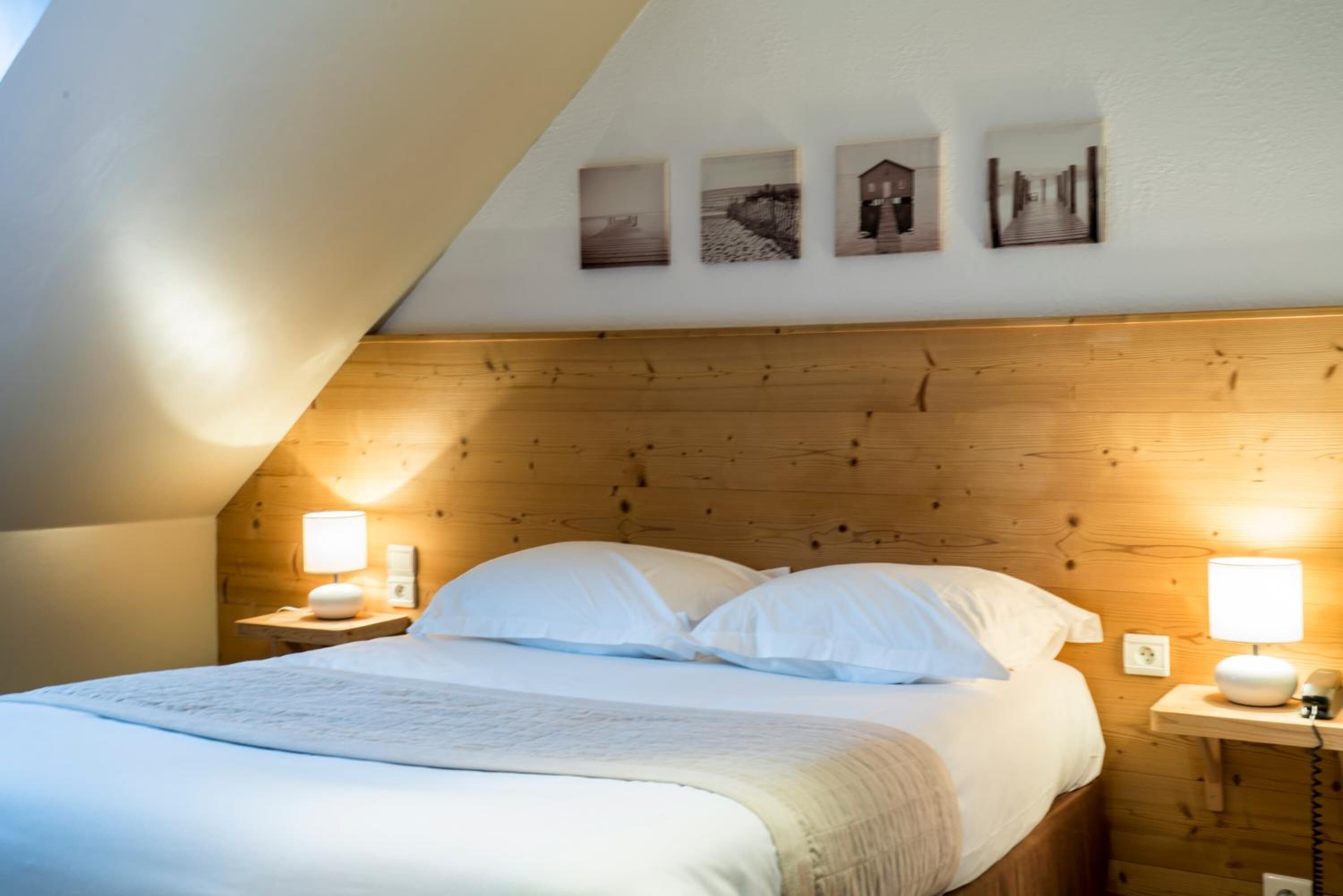 A bedroom with a wooden headboard, a bed with white linens, two bedside lamps, and three framed pictures on the wall.